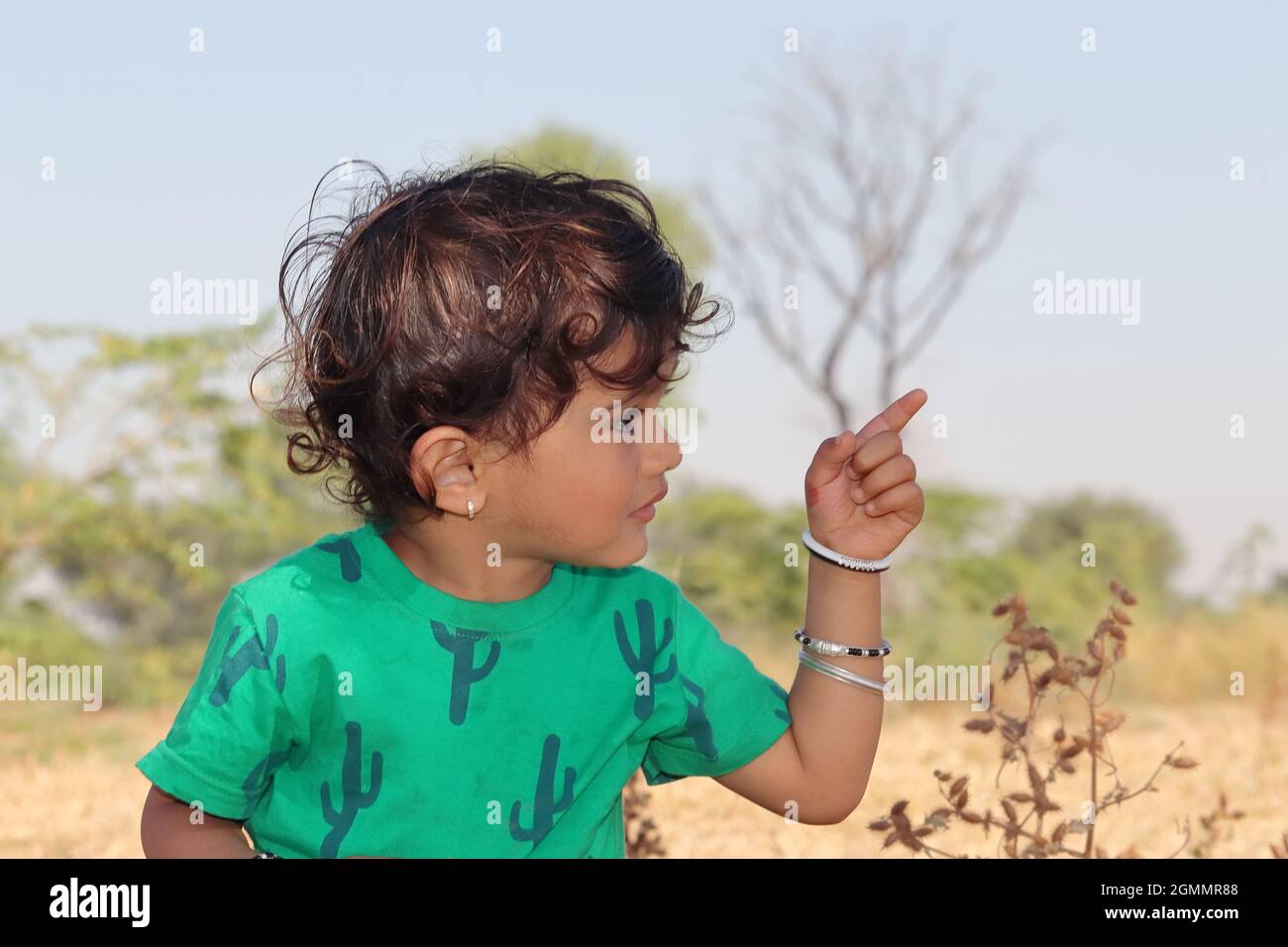 Close-up profile view portrait of A small cute Indian Hindu child wearing a green shirt stands outside, showing a distant object with a finger gesture Stock Photo