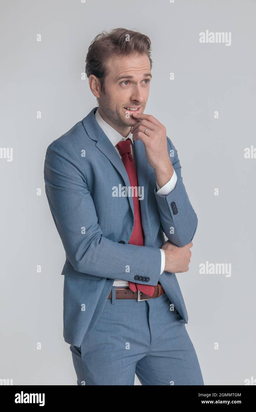 thoughtful young man in blue suit holding hand to chin and thinking, dreaming and smiling while folding arm and posing in front of grey background in Stock Photo