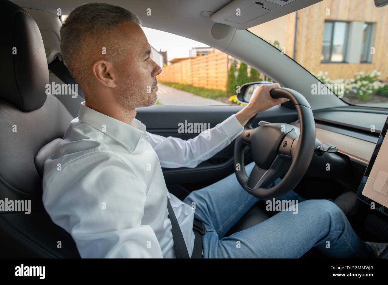Serious mature man in jeans and white shirt sitting by steering wheel while driving new electric car Stock Photo