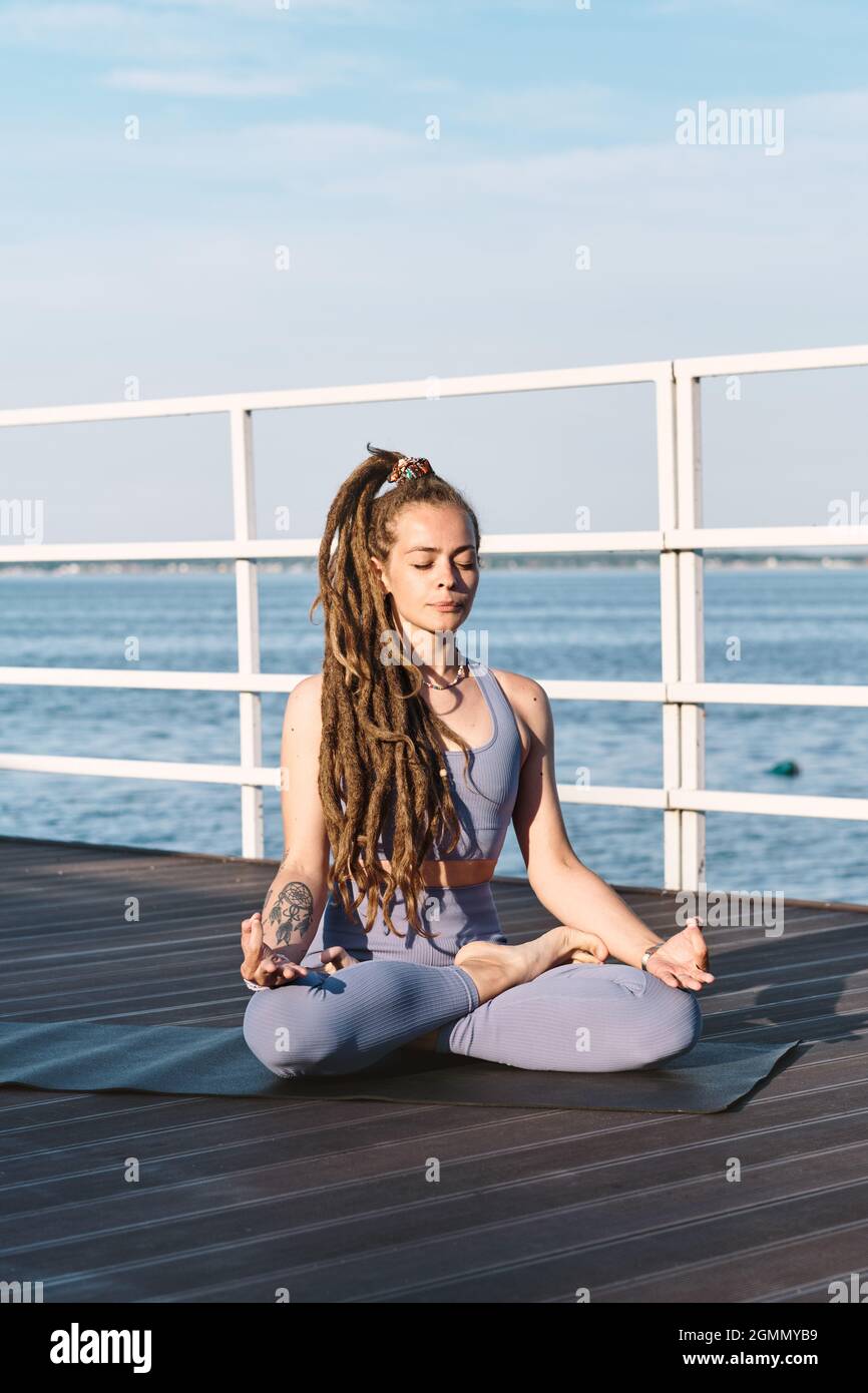 Contemporary sportswoman sitting in pose of lotus on pier against blue sky and water Stock Photo