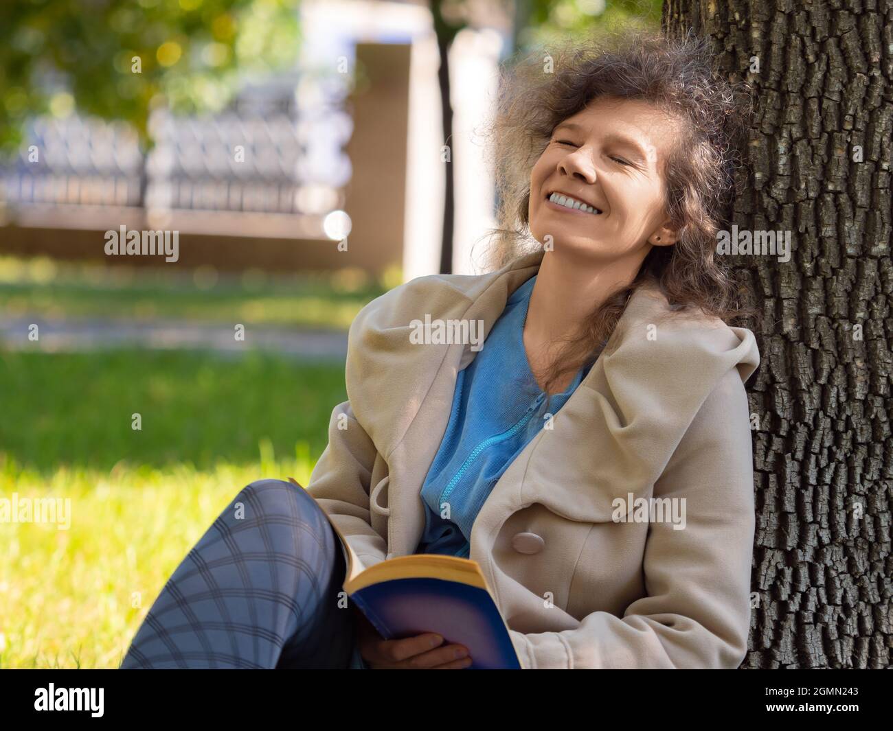 Photo of a middle-aged woman with a book in the park. She sitting on the grass in city park with a book and smiling with closed eyes. Concept of rest Stock Photo