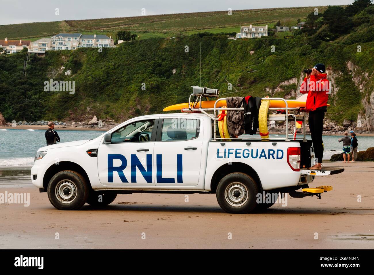 A lifeguard watches the swimmers through binoculars from a RNLI patrol vehicle parked on Bantham Beach, Devon. Stock Photo