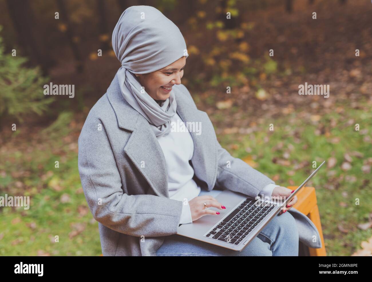Young asian muslim woman using laptop at autumn park. Online education ...