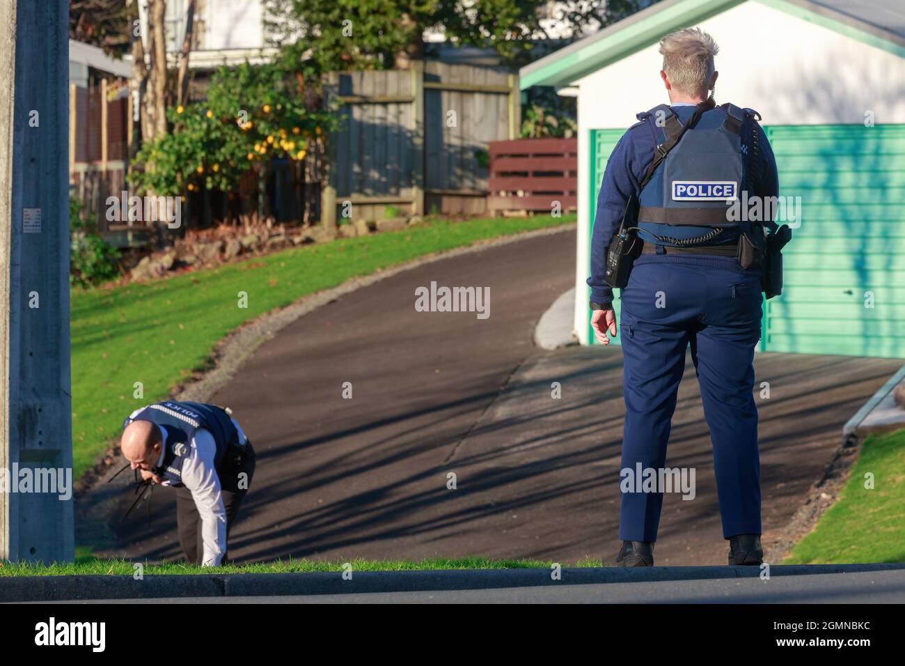 Members of the New Zealand police force in a suburban area. One stands guard while another searches the ground Stock Photo
