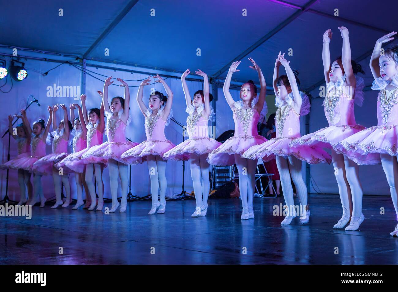 A row of young Asian girl ballerinas on stage in pink tutus, standing in the 'releve' position with arms raised Stock Photo