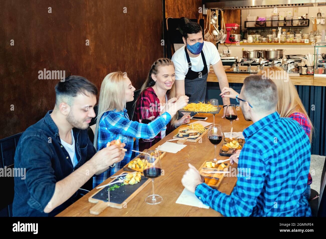 Happy best friends enjoy time together at restaurant while waiter wearing face mask serving food during covid pandemic after business reopening Stock Photo