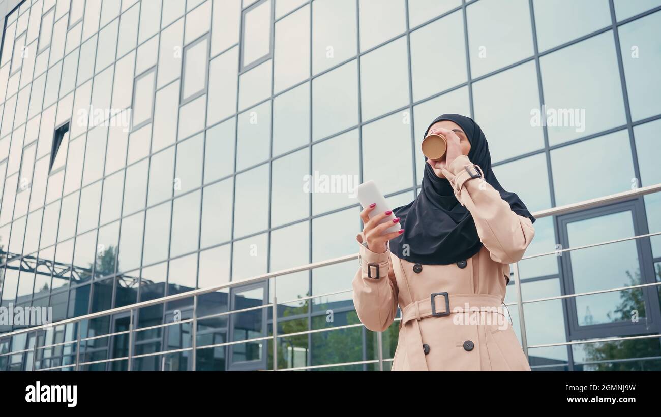 Young muslim woman in hijab holding smartphone and drinking coffee to go near building Stock Photo
