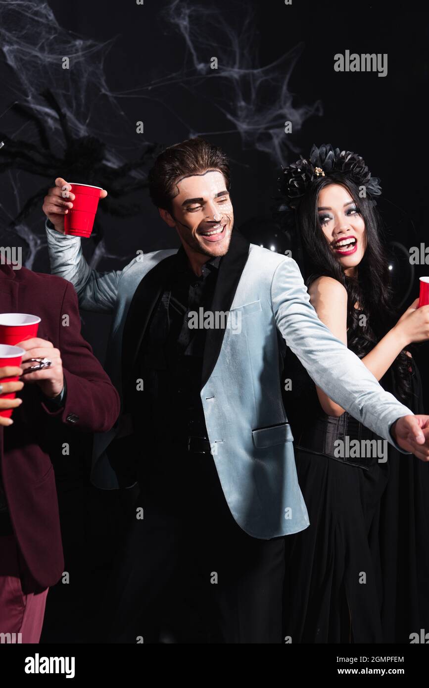 happy interracial friends holding plastic cups while dancing on halloween party on black Stock Photo