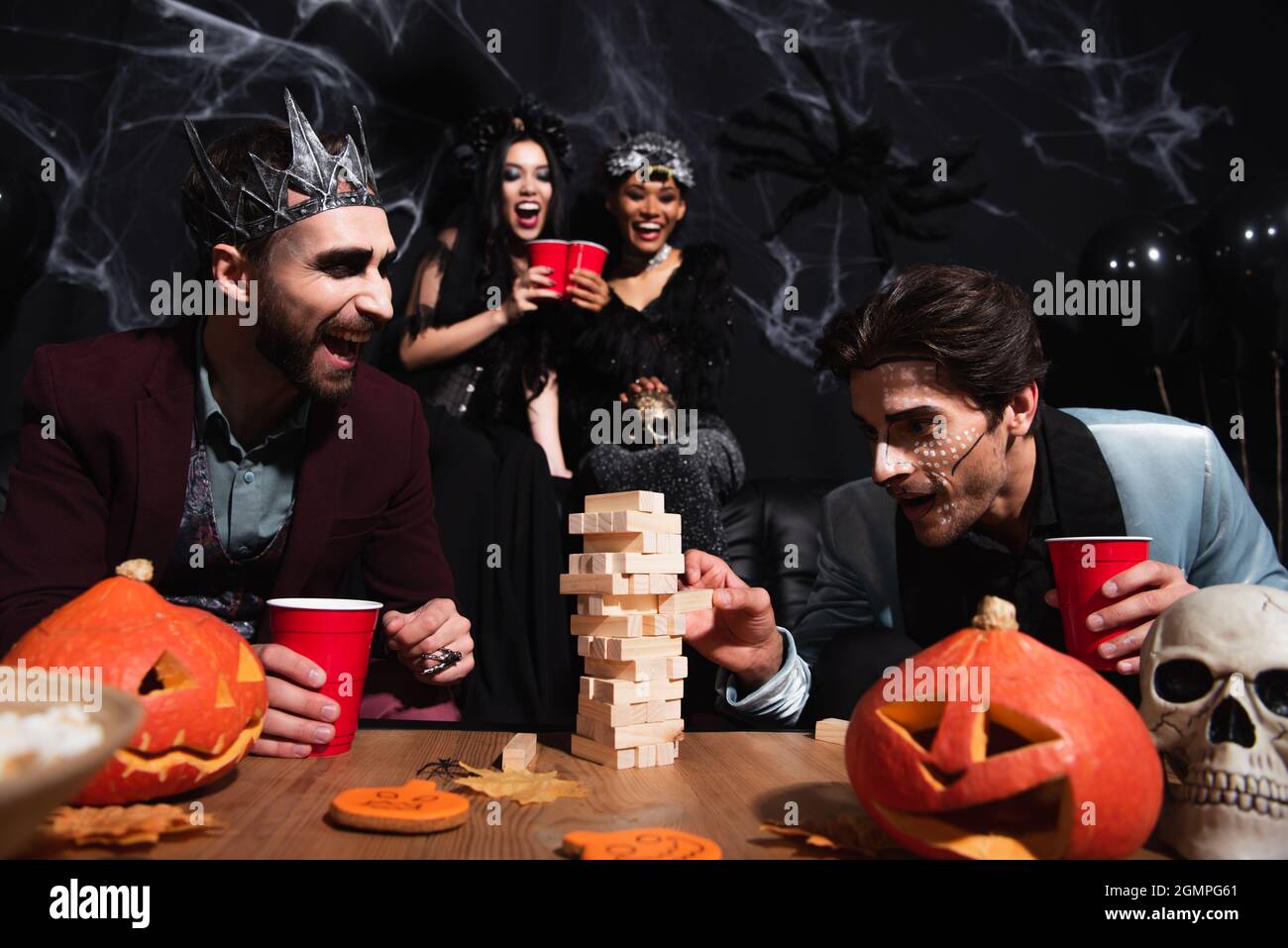 man in halloween makeup playing wood blocks game while blurred interracial women toasting with plastic cups on black Stock Photo