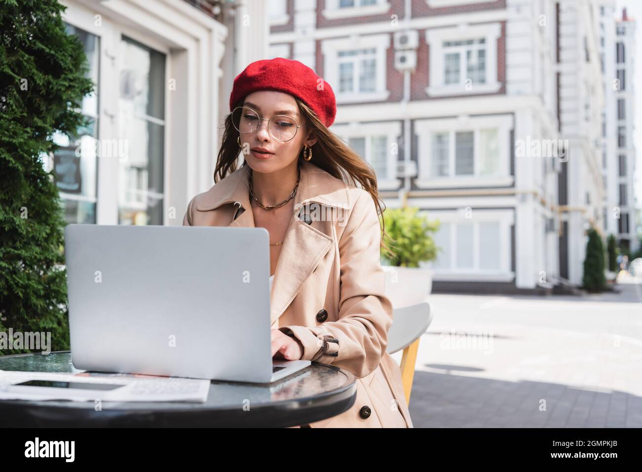 pretty freelancer in eyeglasses, red beret and beige trench coat using laptop on terrace of cafe Stock Photo