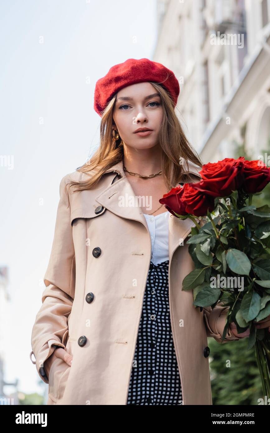 elegant woman in trench coat and red beret holding roses and posing with  hand in pocket outside Stock Photo - Alamy