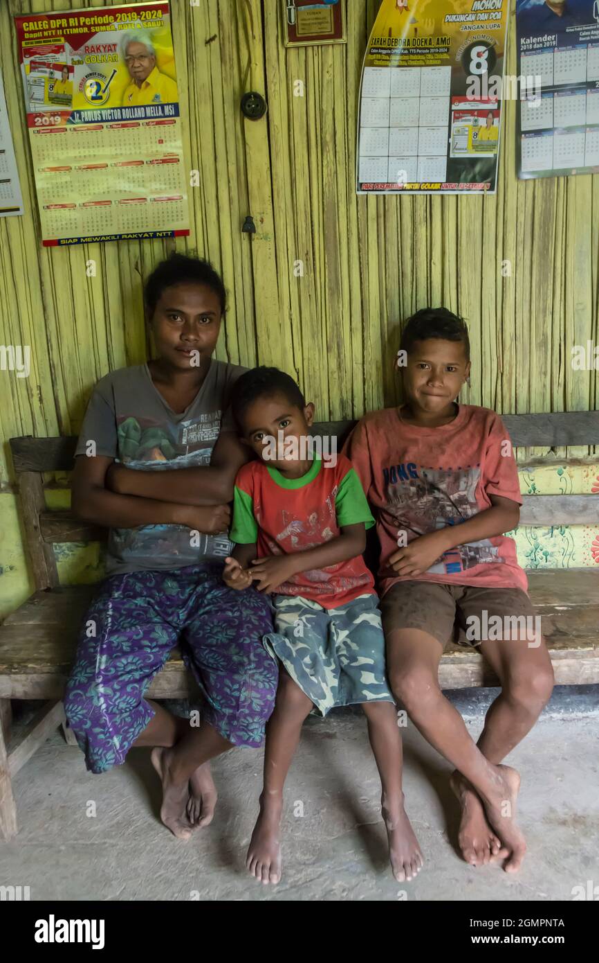 Mother and two sons sitting on a wooden bench against a yellow painted bamboo wall in Oinlasi village, West Timor, Indonesia. Stock Photo
