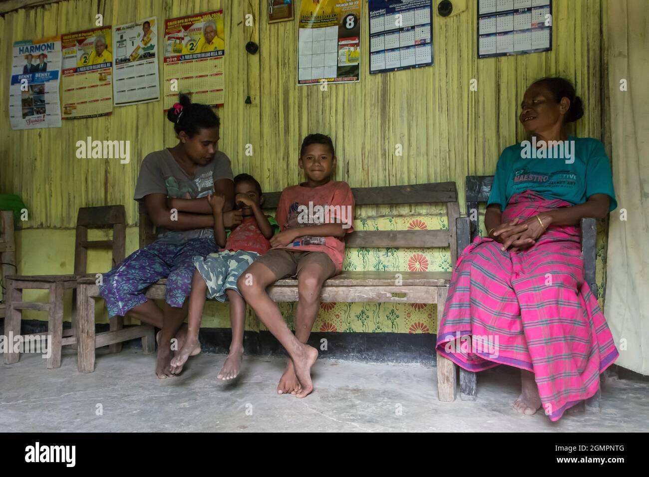 Mother,sons and grandmother sitting on a wooden bench in a room with yellow painted bamboo walls. Oilasi village, West Timor, Indonesia Stock Photo
