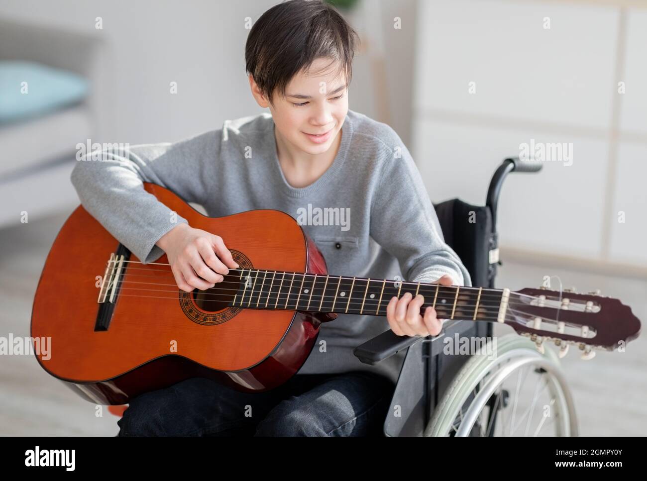 Cheerful impaired teen boy in wheelchair learning how to play guitar and singing indoors Stock Photo