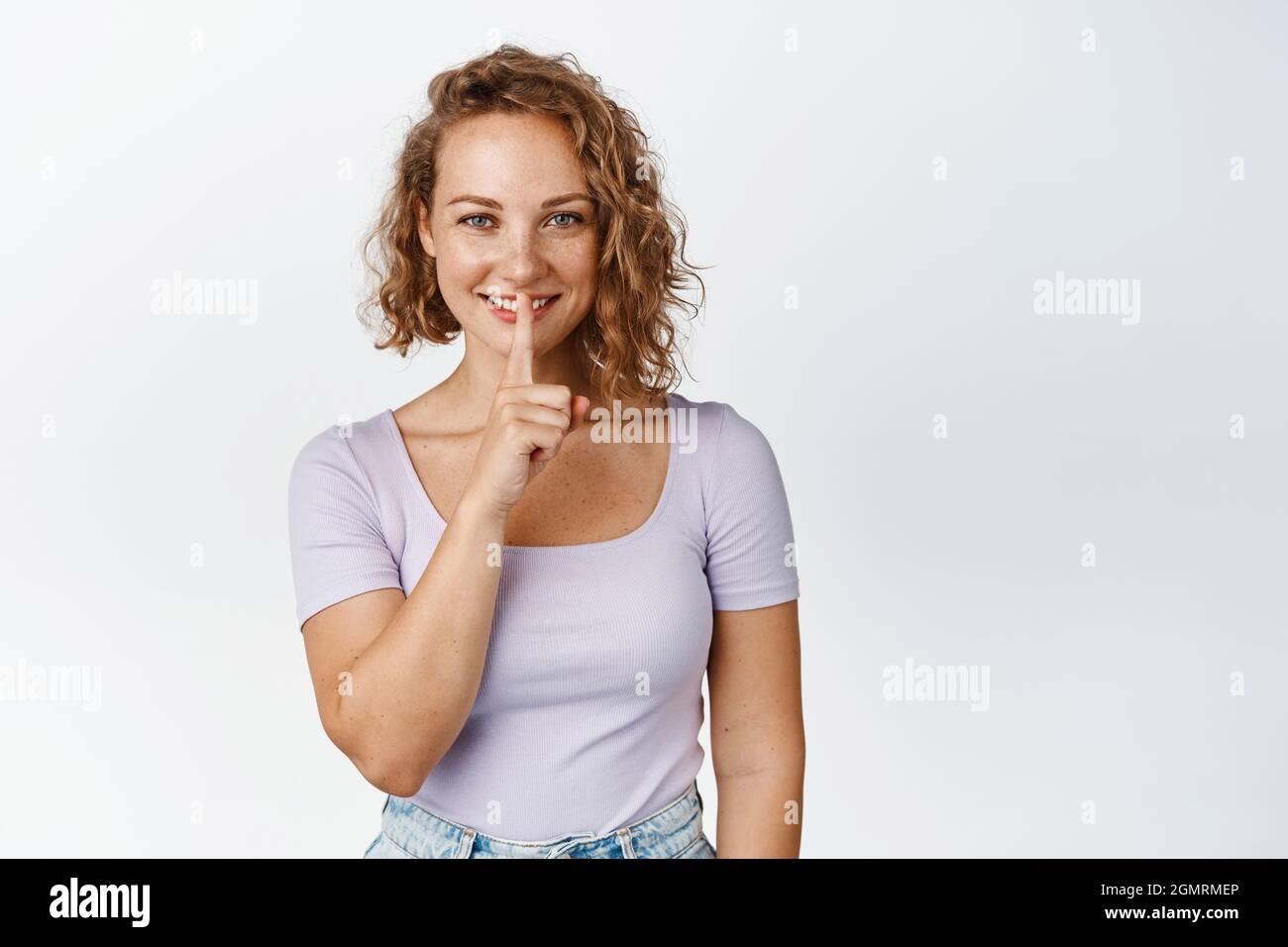 Smiling blond woman make hush sign, shushing with finger on her lips, be quiet gesture, standing in tshirt over white background Stock Photo