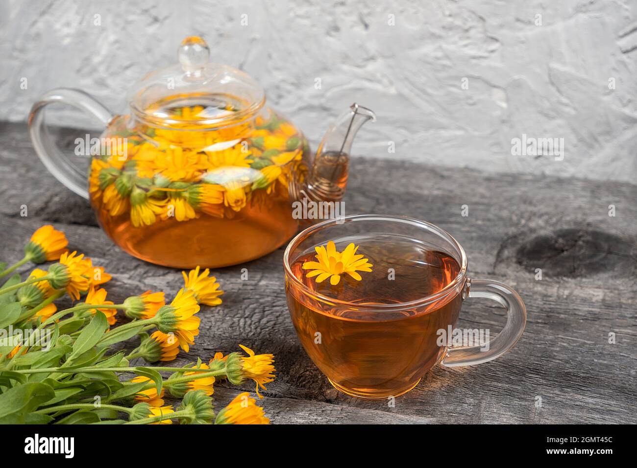 Cup of herbal tea and transparent teapot and marigold flowers on wood
