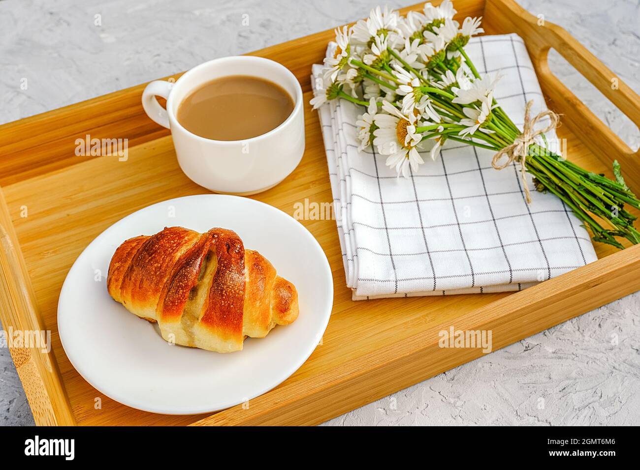 Cup of coffee with milk, freshly baked croissant, checkered napkin and camomile flowers on wooden tray on gray stone table. Concept Good morning. Stock Photo