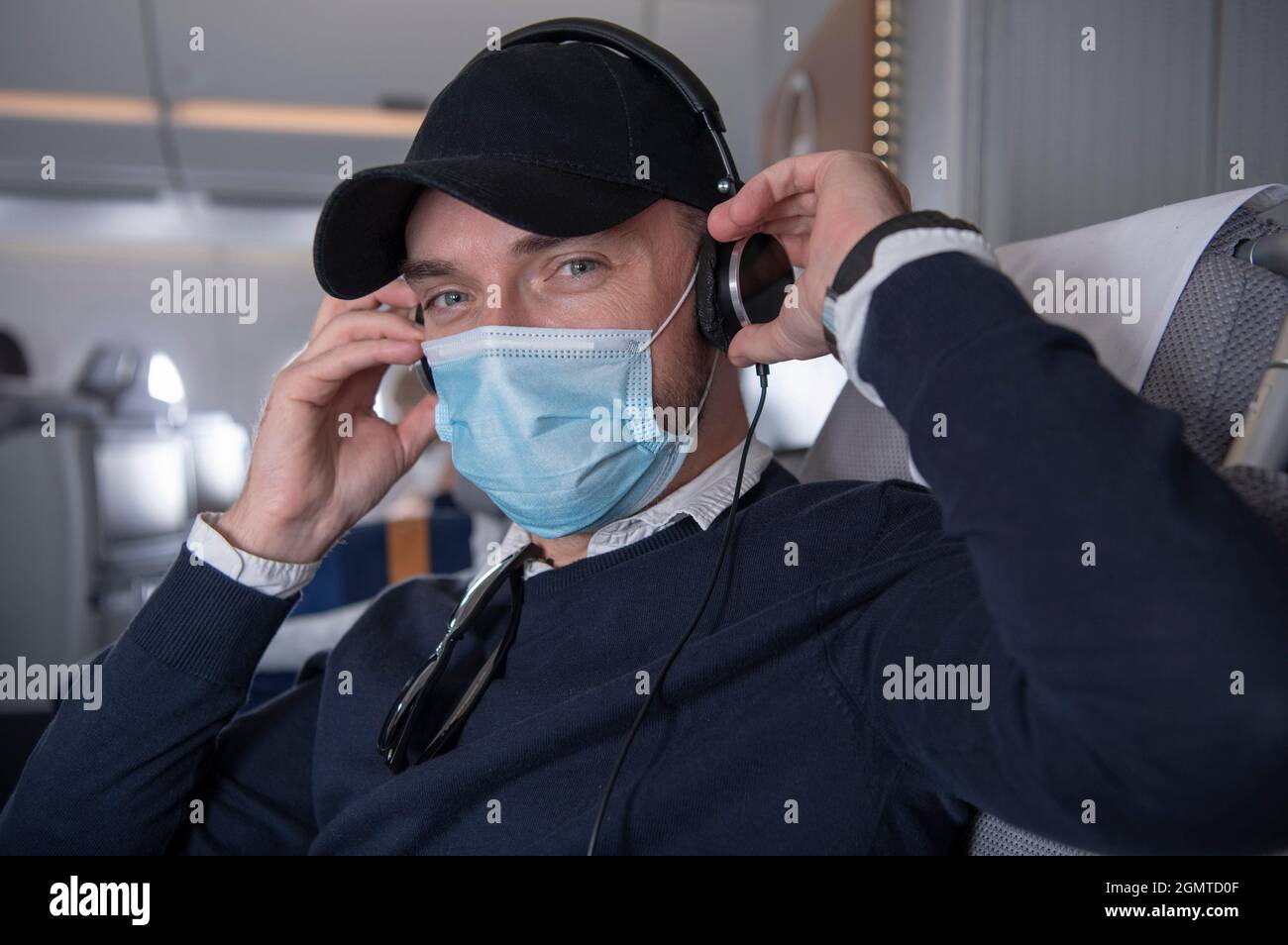Happy Caucasian Business Class Flight Passenger in His 40s Wearing Mask with Headphones Listen Music and Watch Movies on Board of an Airplane. Stock Photo