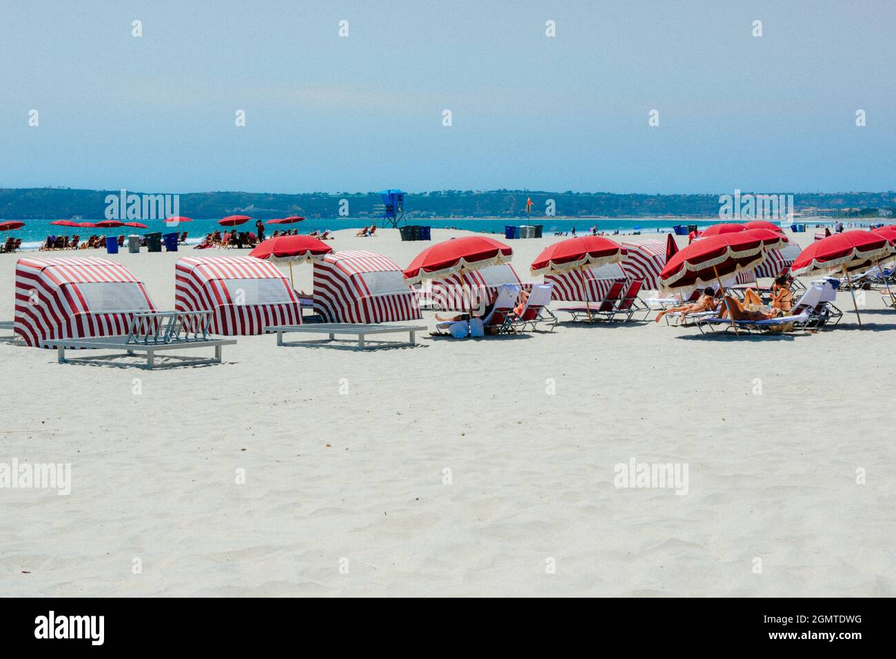 White sandy beach with beach sunshade recliners, people, bikinis,  blue water, red stripes and Victorian fringed umbrellas, Coronado Island, CA Stock Photo