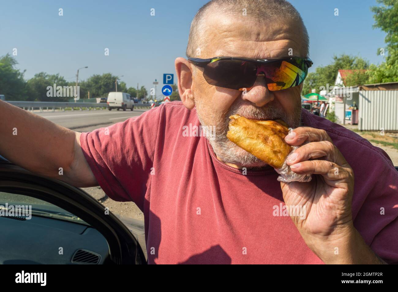 Portarait of hungry Cacasiam senior driver wearing sunglasses eating patty near his car standing on Ukrainian road Stock Photo