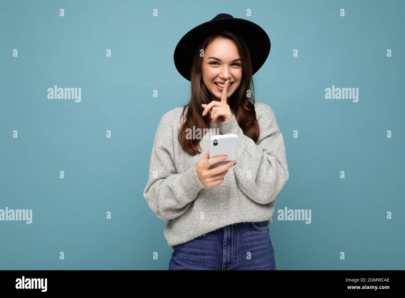 Attractive young smiling woman wearing black hat and grey sweater holding smartphone looking at camera showing shhh gesture isolated on background Stock Photo