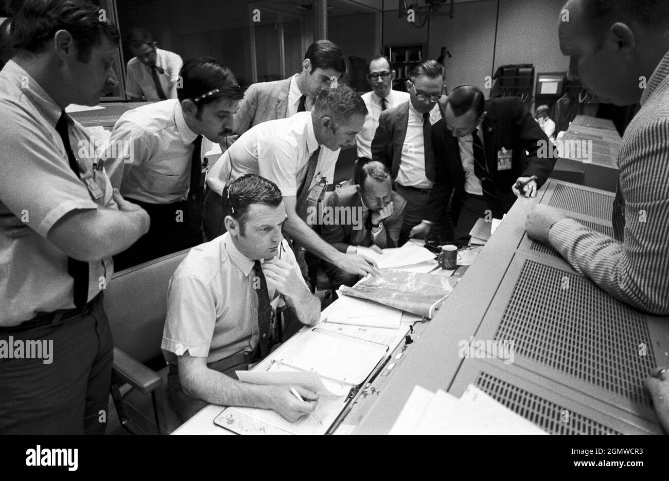 (15 April 1970) --- A group of flight controllers gathers around the console of Glenn S. Lunney (seated, nearest camera), Shift 4 flight director, in the Mission Operations Control Room (MOCR) of Mission Control Center (MCC), located in Building 30 at the Manned Spacecraft Center (MSC). Their attention is drawn to a weather map of the proposed landing site in the South Pacific Ocean. Among those looking on is Dr. Christopher C. Kraft, deputy director, MSC, standing in black suit, on right. When this photograph was taken, the Apollo 13 lunar landing mission had been canceled, and the problem-pl Stock Photo