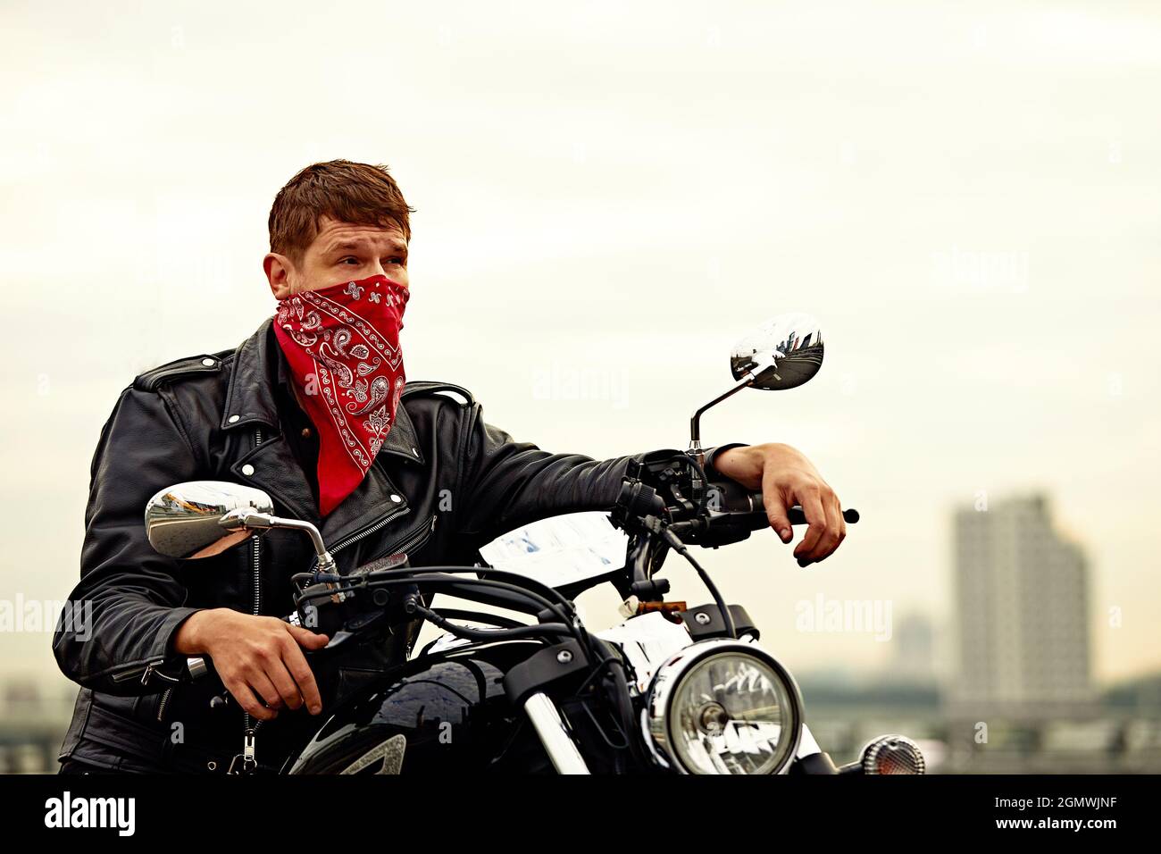 Portrait of stylish male dressed in a black leather jacket and bandana  sitting on his custom-made retro motorcycle, looking away, buildings of the  big Stock Photo - Alamy