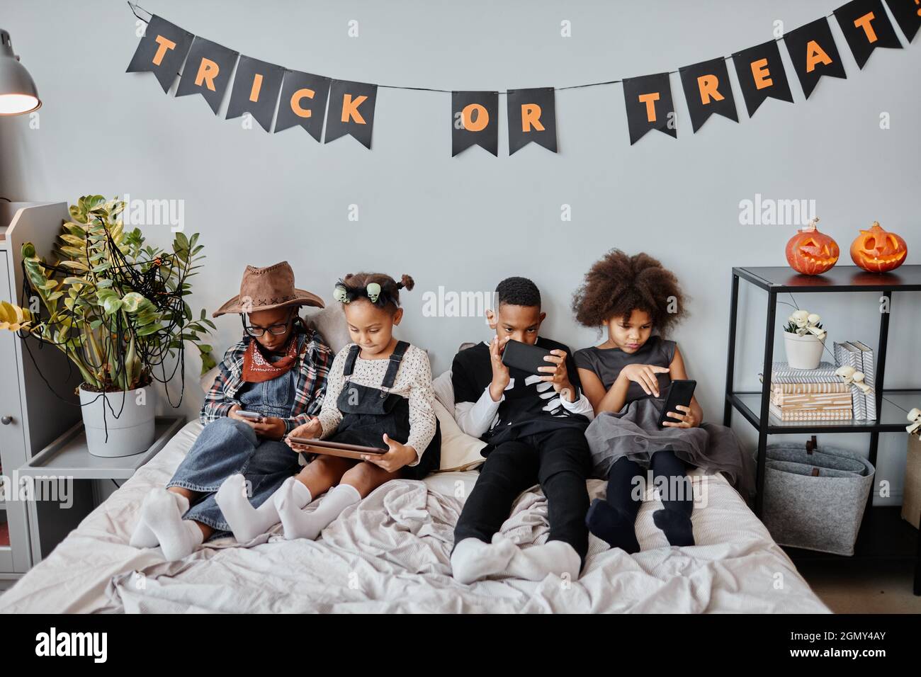 High angle portrait of four African-American children using gadgets in bed under Halloween decorations at home Stock Photo