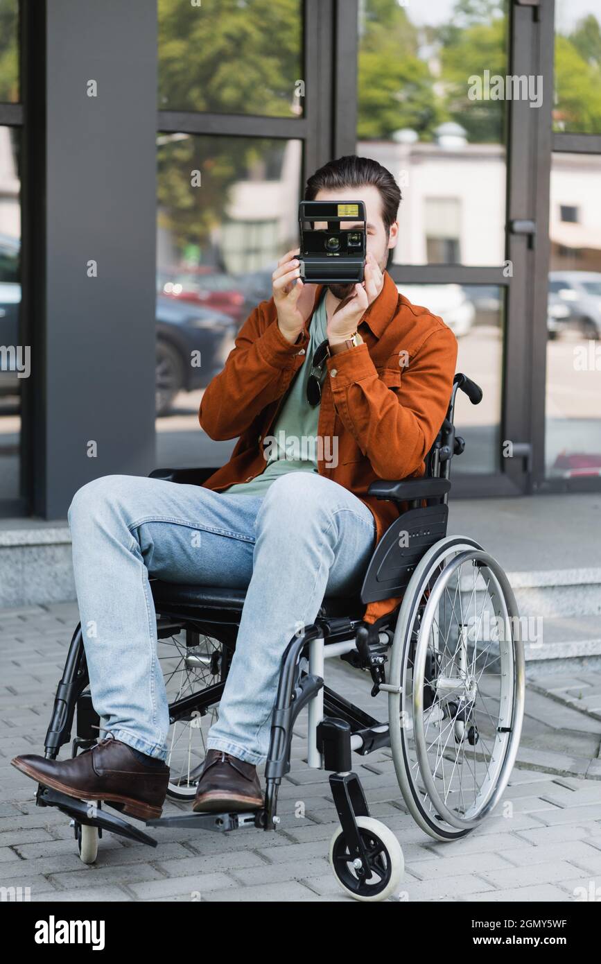 full length view of disabled man in wheelchair taking picture on vintage camera on street Stock Photo