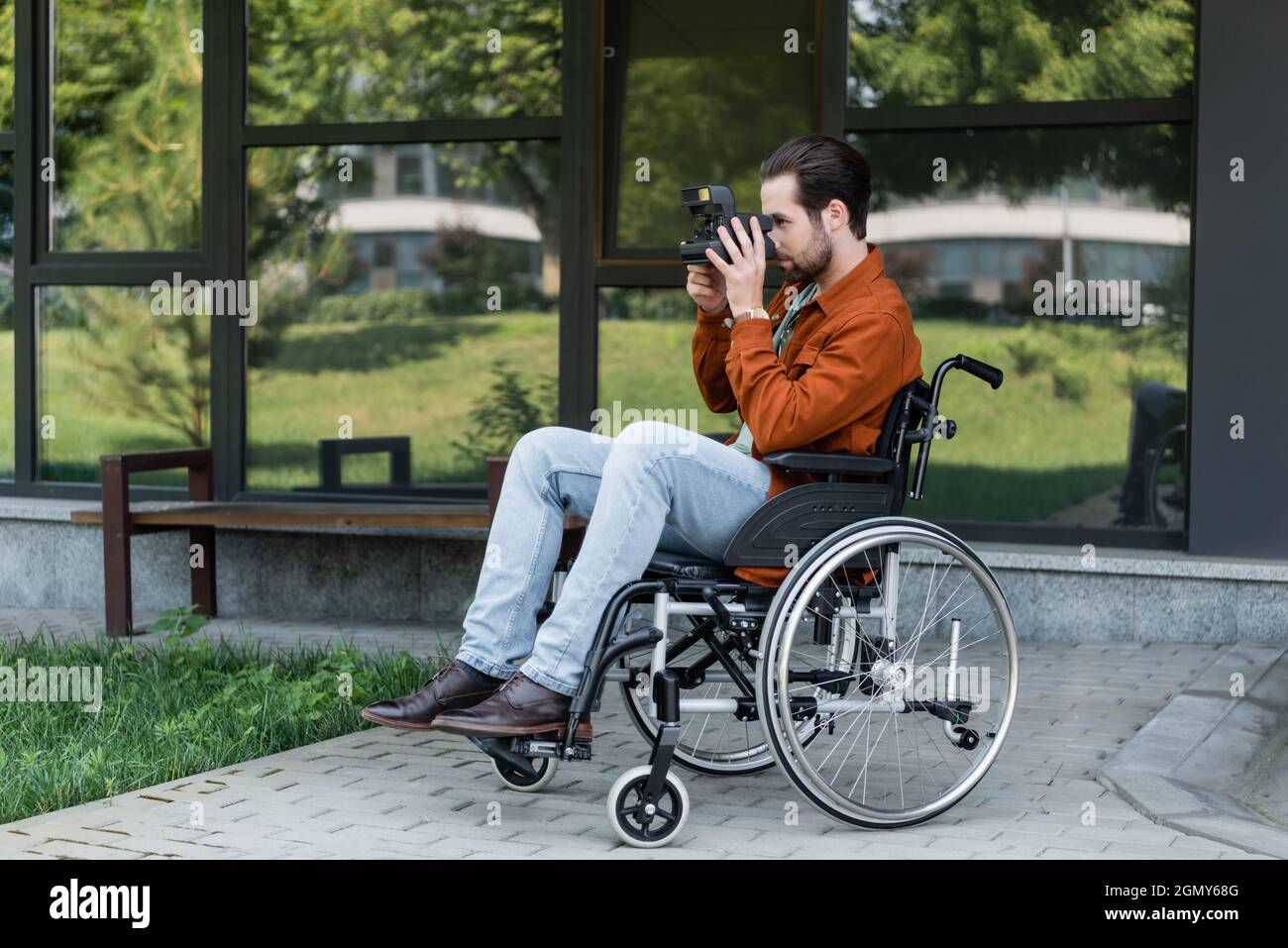 young disabled man in wheelchair taking photo on vintage camera near building with glass facade Stock Photo