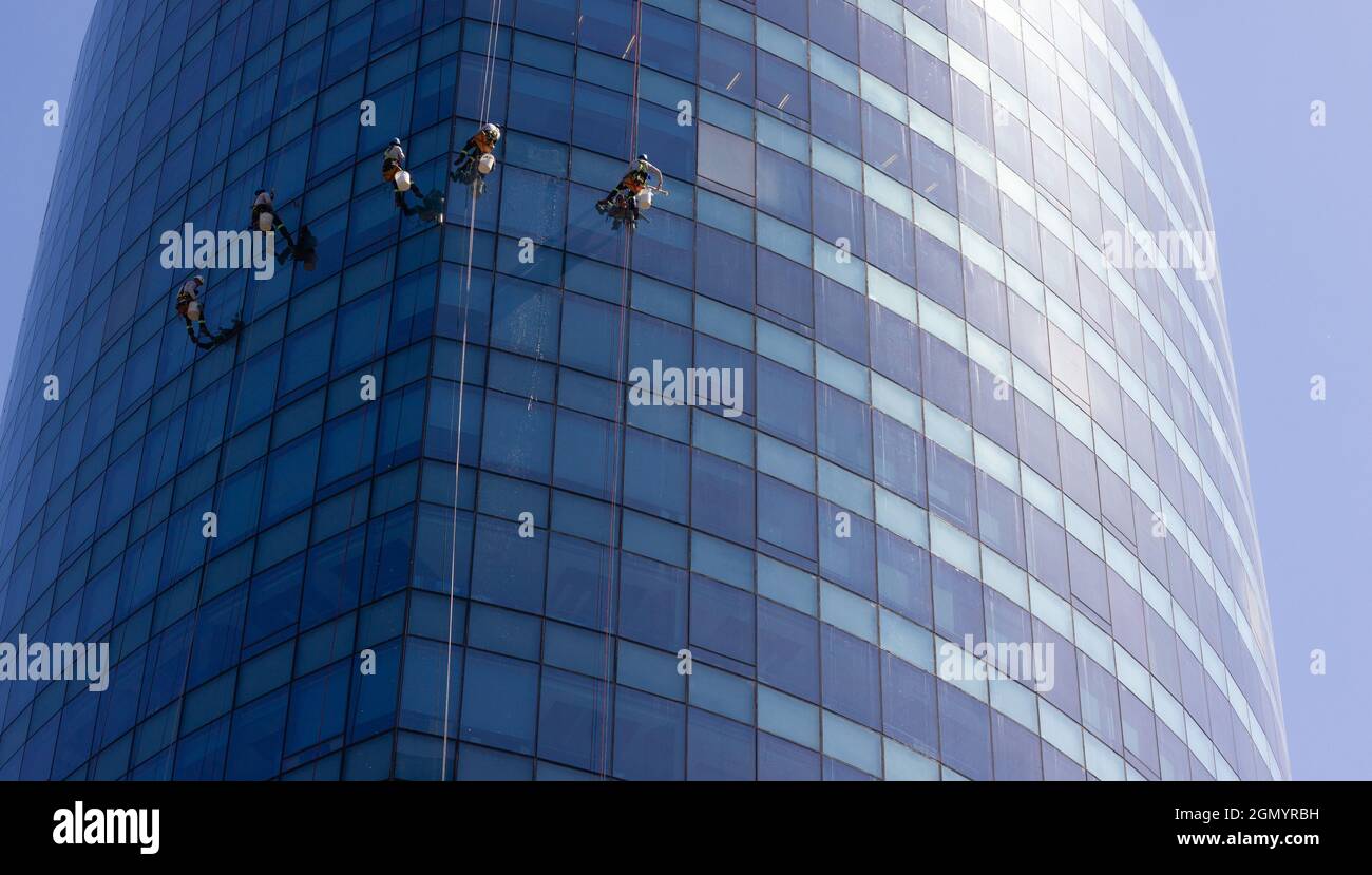 Five window washers hanging with ropes outside blue glass modern building. Risky job, dangerous work concepts Stock Photo