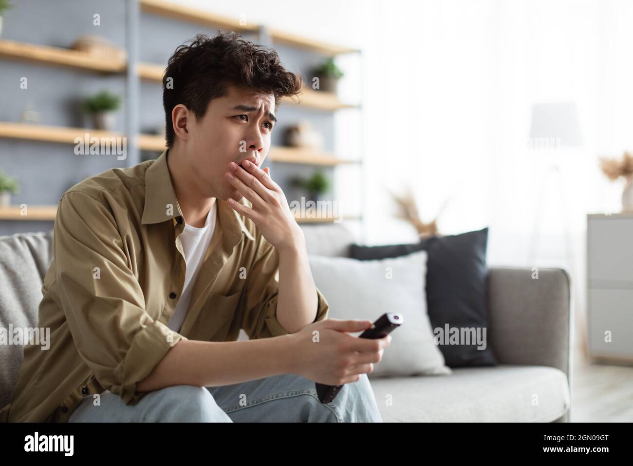 Bored asian man watching television, yawning sitting on couch Stock Photo
