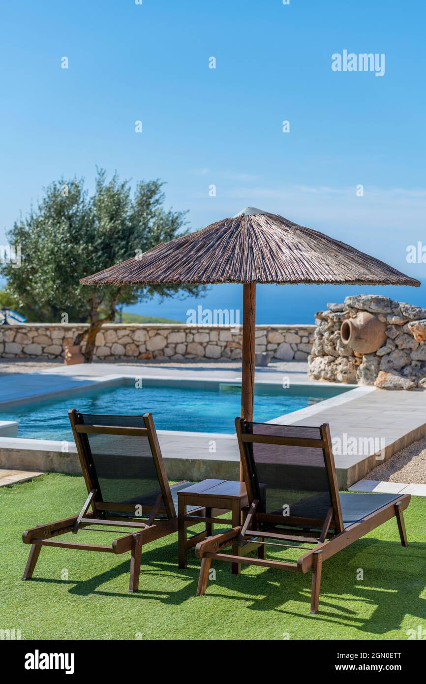 two sun beds or sun loungers under a raffia or wicker tropical style umbrella at the side of a swimming pool at a private holiday villa on zakynthos. Stock Photo
