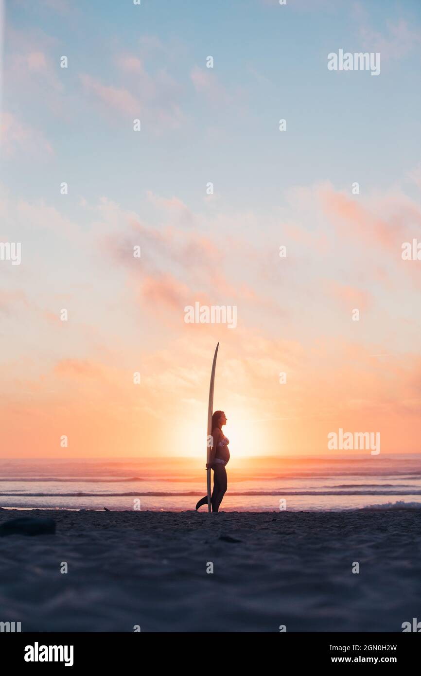pregnant woman standing with surfboard on the beach, surfing, pregnancy