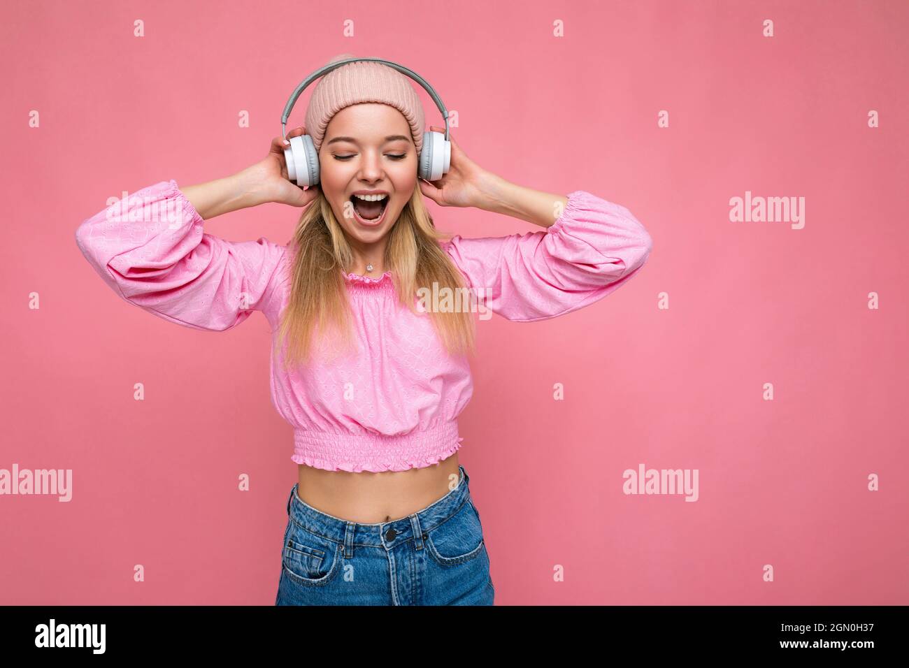 Attractive positive emotional smiling young blonde woman wearing pink blouse  and pink hat isolated over pink background wall wearing white wireless  Stock Photo - Alamy