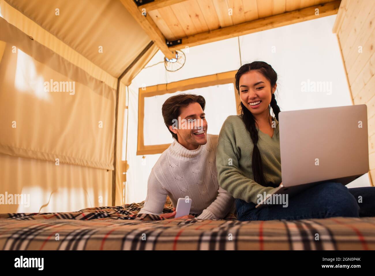 Multiethnic couple using gadgets on bed in glamping house Stock Photo