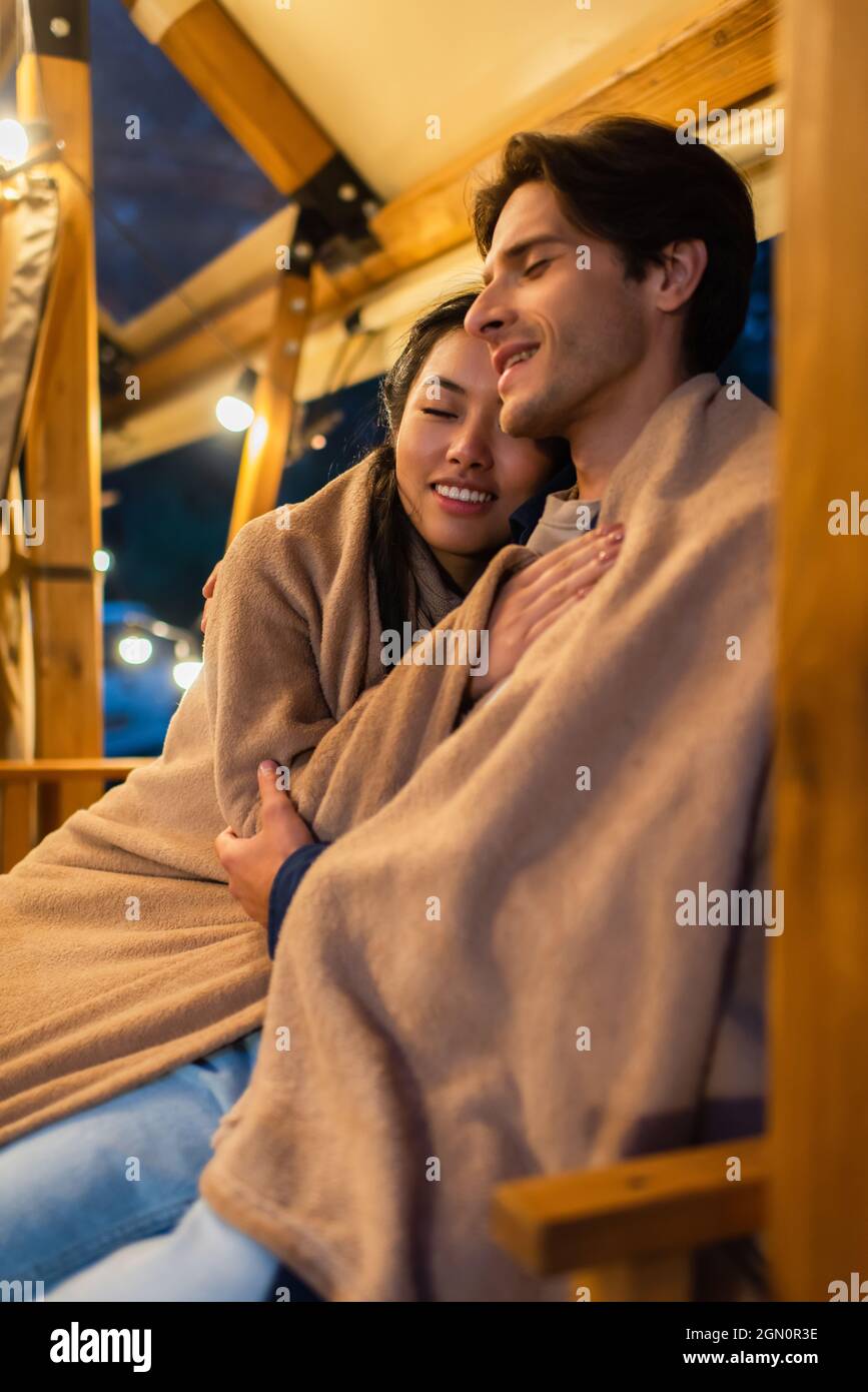 Smiling asian woman in blanket hugging boyfriend on glamping terrace ...