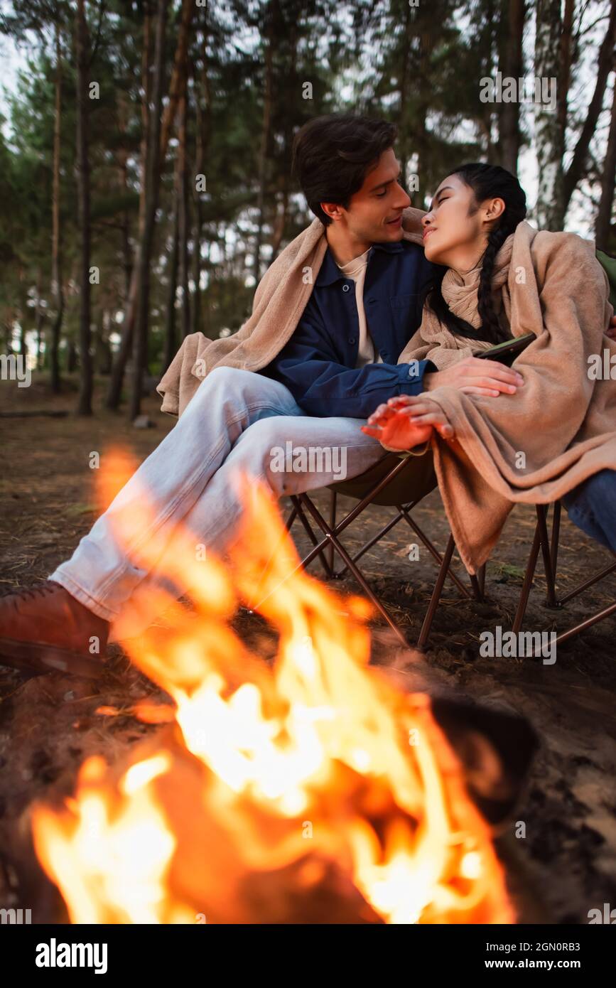 Interracial couple in blankets kissing near blurred campfire Stock