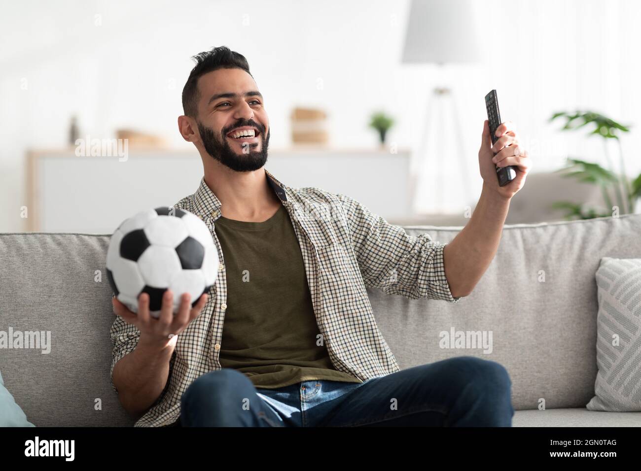 Positive young Arab guy watching soccer game on TV, holding remote control and ball at home Stock Photo