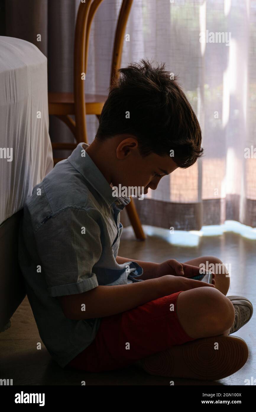 8 years old child sitting on the floor and playing with his smartphone. In the background, curtain being illuminated by natural light. Stock Photo
