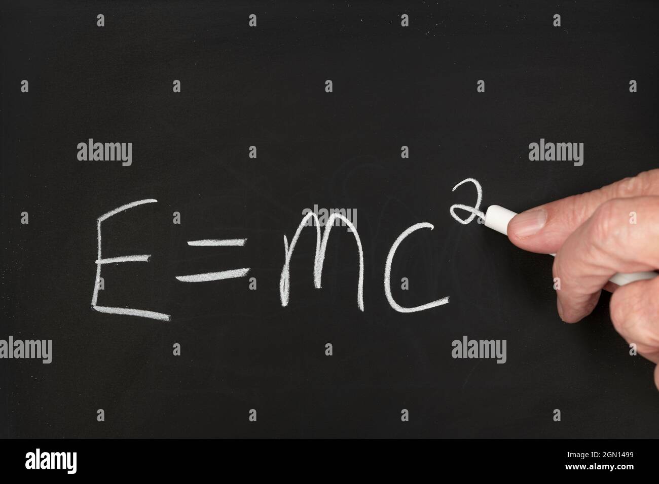 A teacher writes the theory of relativity Einstein equation on a black chalkboard while teaching a group of students in a classroom. Stock Photo