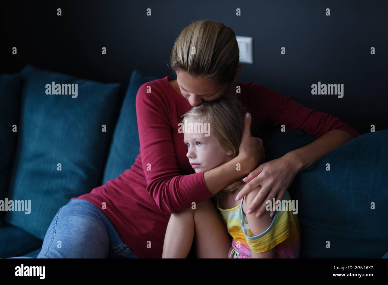 Woman and sad little daughter are sitting on couch and hugging Stock Photo