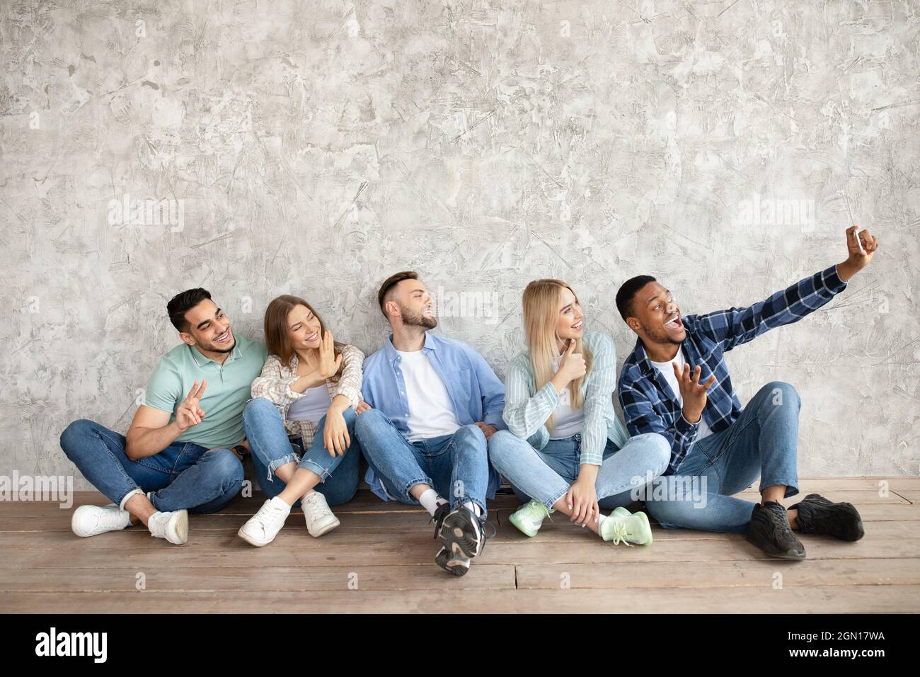 Full length of young diverse students sitting near grey studio wall, taking selfie with cellphone, copy space Stock Photo