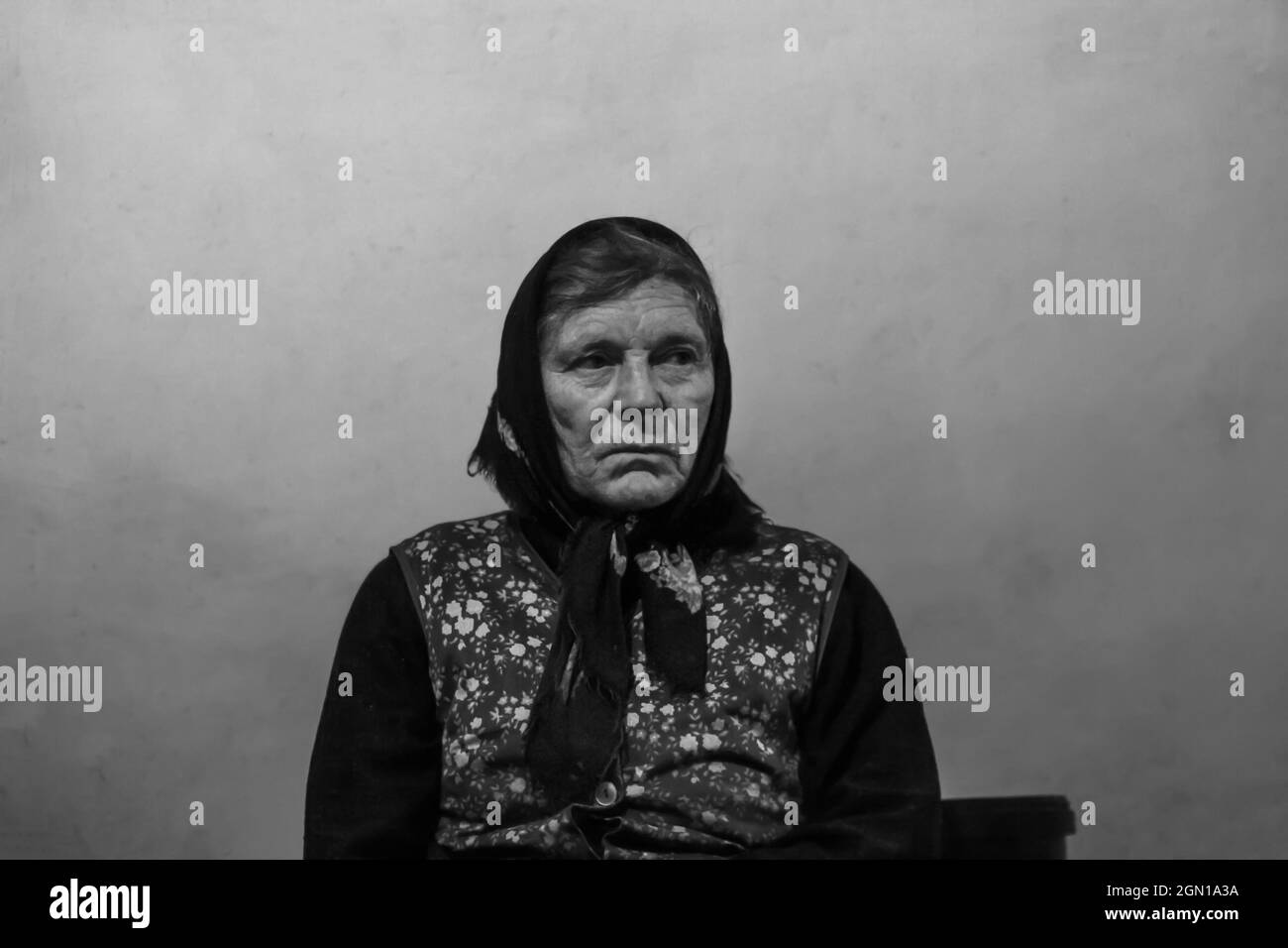 Defocus portrait of 60s russian grandmother senior old woman seating indoors. Old women shawl. Sad person. White dirty background. Black and white. Ou Stock Photo