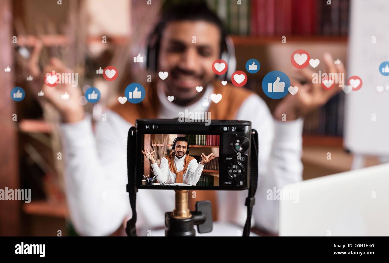 Smiling millennial arab guy recording video blog at home, double exposure with likes and icons Stock Photo