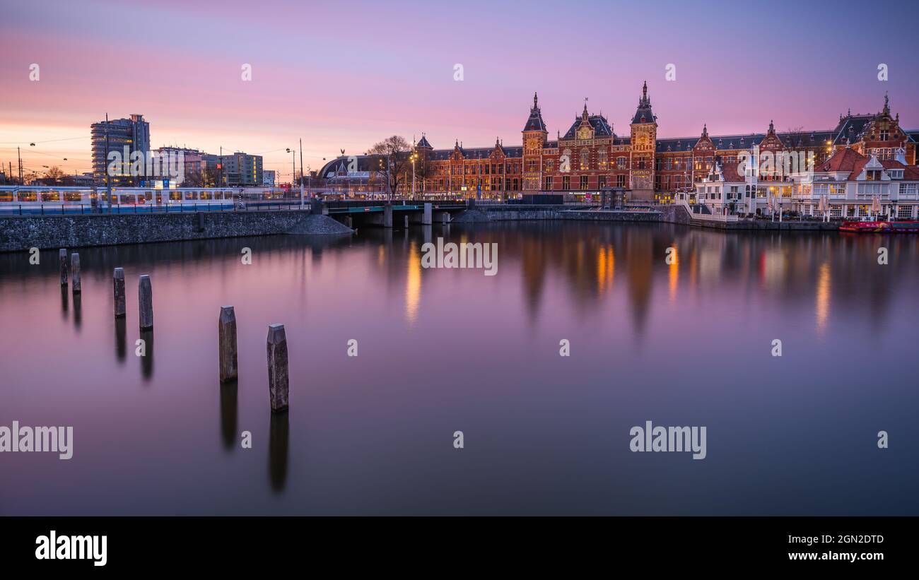 NETHERLANDS, AMSTERDAM, CENTRAL STATION AT SUNSET WITH THE CANAL IN THE FOREGROUND Stock Photo