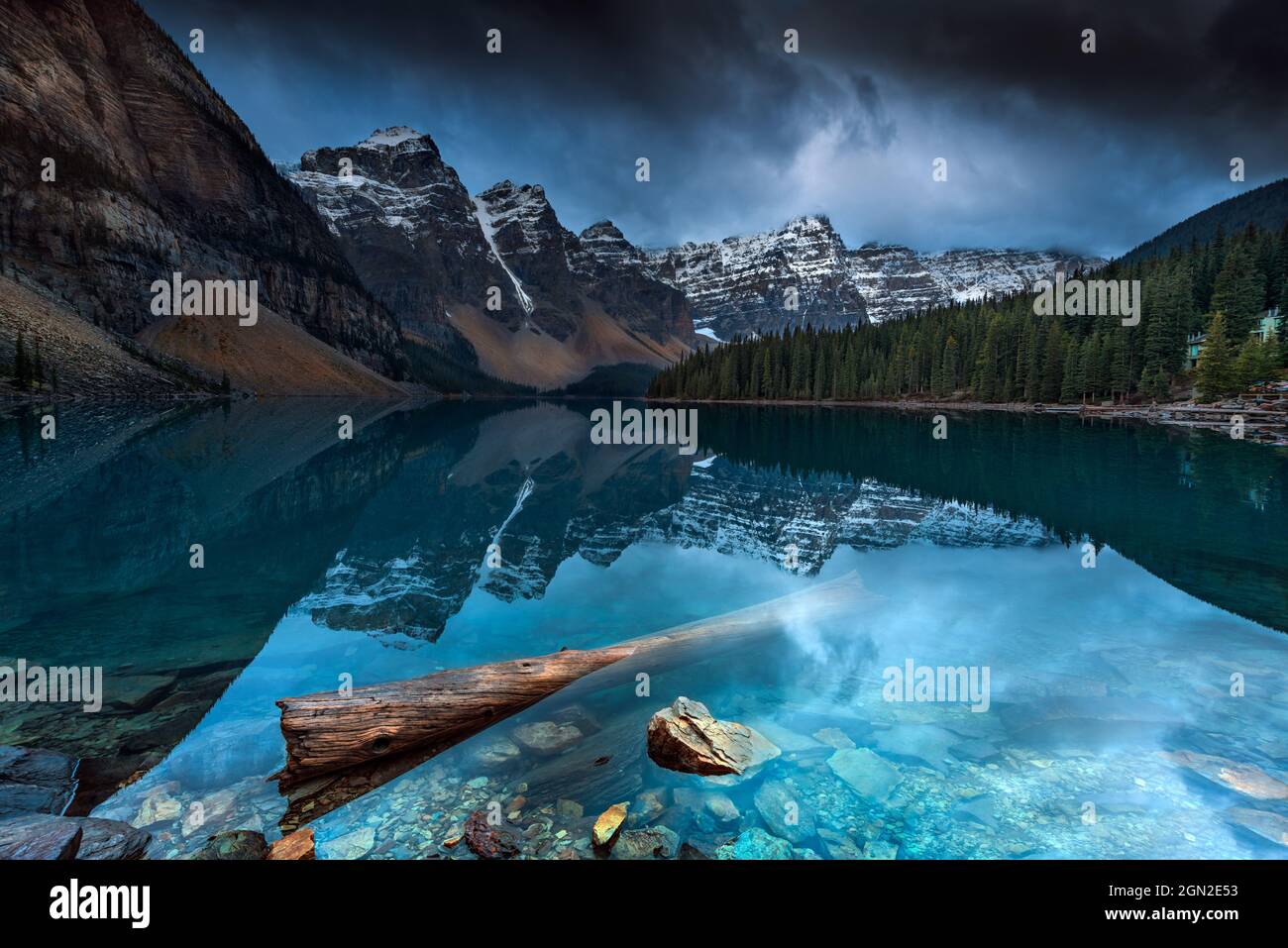 CANADA, ALBERTA, BANFF PARK, MORAINE LAKE. TREE TRUNK FLOATING IN THE LAKE WITH A VIEW OF THE MAJESTIC PEAKS OF THE CANADIAN ROCKIES UNDER A STORMY SK Stock Photo