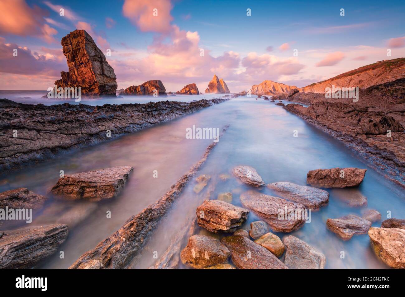 SPAIN, CANTABRIA, LIENCRES. VIEW FROM BELOW OF ARNIA BEACH WITH ITS ROCKS CUT OUT AT SUNSET AND IN THE FOREGROUND A PILE OF PEBBLES IN THE WATER Stock Photo