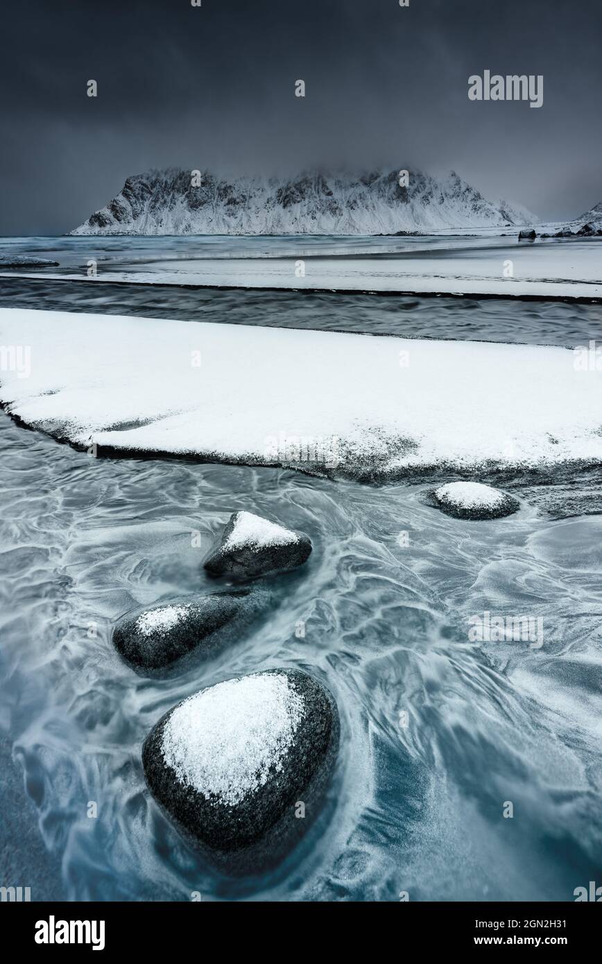 NORWAY, LOFOTEN ISLANDS, FLAKSTAD. FLAKSTAD BEACH IN WINTER WITH SMALL SNOWY STONES IN THE FOREGROUND IN THE WATER AND THE MOUNTAINS IN THE DISTANCE U Stock Photo