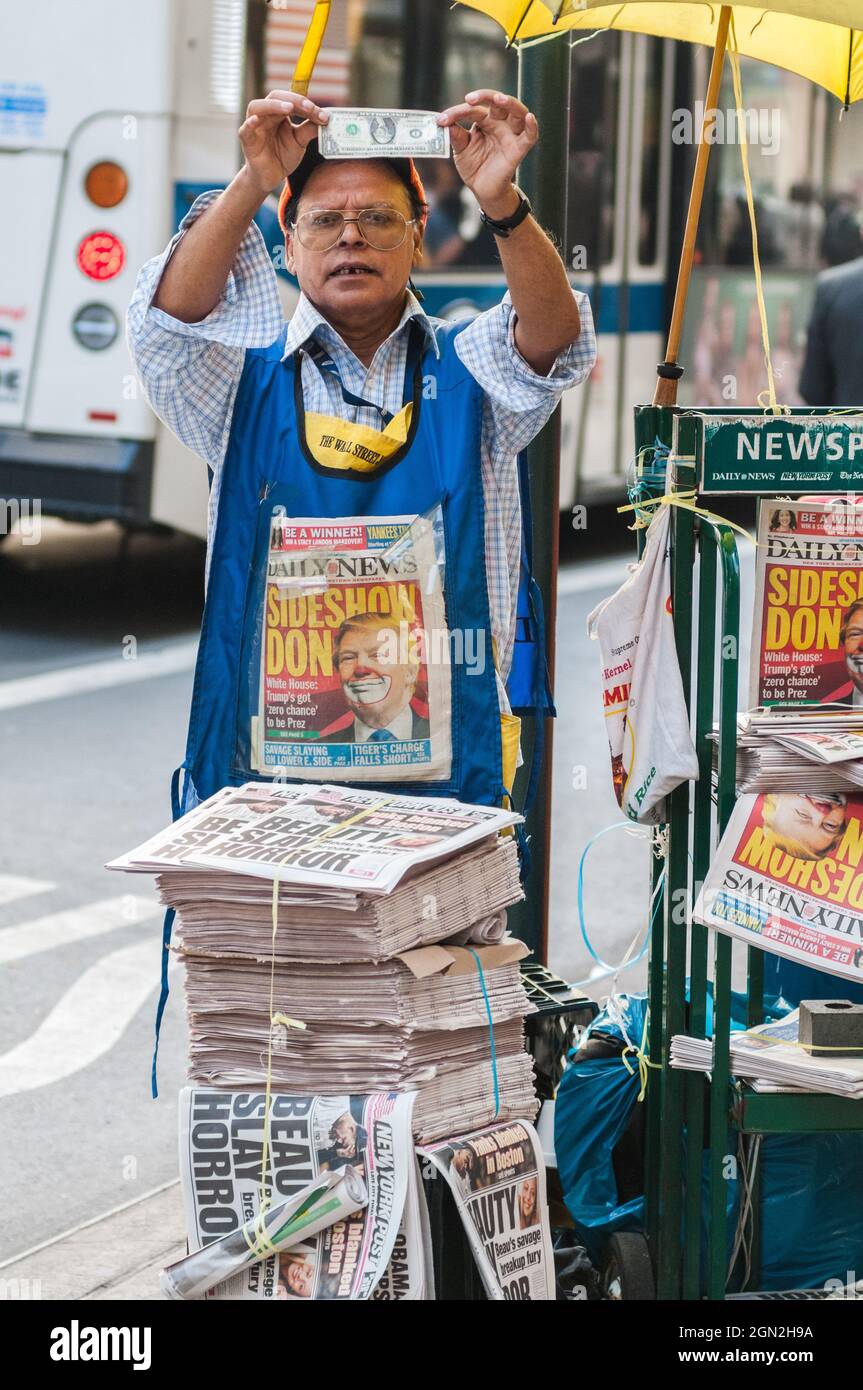 UNITED STATES, NEW YORK, MANHATTAN, A NEWSPAPER SALESMAN WITH A FRONT PAGE OF TRUMP IN CLOWN SHOWING ME A US DOLLAR Stock Photo